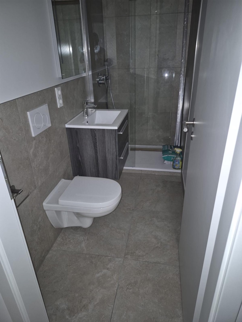Schmales Duschbad mit Natursteinoptik-Fliesen, Wand-WC, Waschtisch und bodenebener Dusche hinter Glas