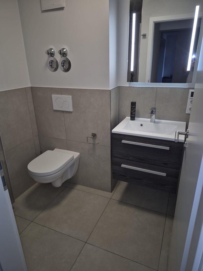 Fertiges Gäste-WC mit grauen Großformatfliesen, Wand-WC, Waschtisch mit dunklem Unterschrank und beleuchtetem Spiegel